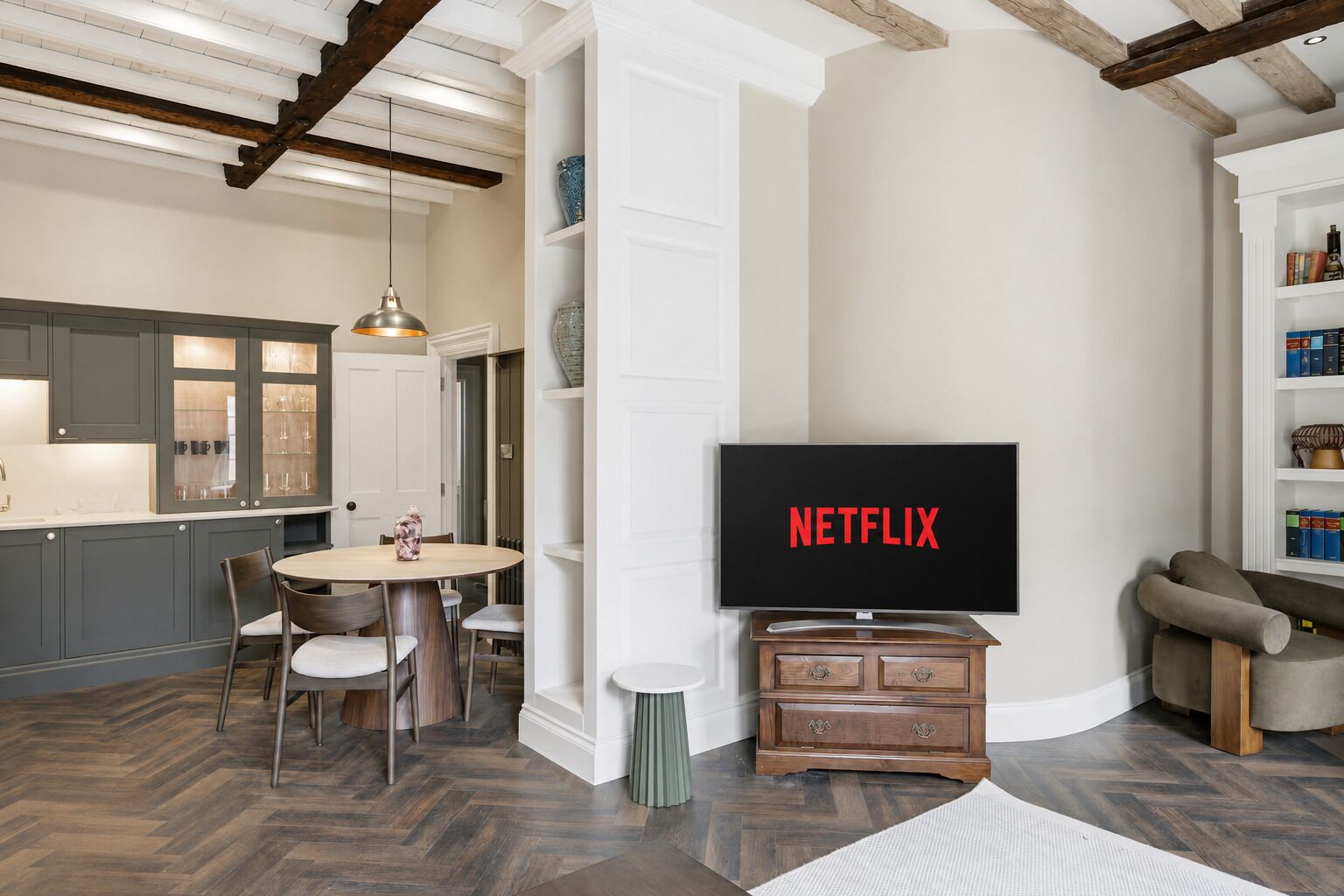 A modern living room with a flat-screen TV displaying 'NETFLIX,' a dining area with a round table and chairs, and a kitchen with built-in cabinets and a pendant light.