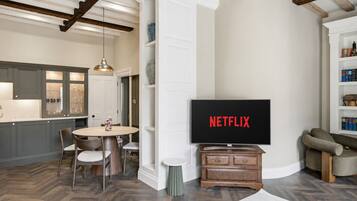 A modern living room with a flat-screen TV displaying 'NETFLIX,' a dining area with a round table and chairs, and a kitchen with built-in cabinets and a pendant light.
