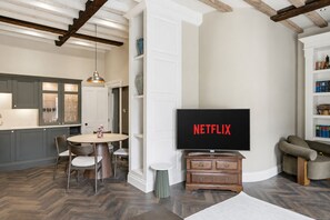 A modern living room with a flat-screen TV displaying 'NETFLIX,' a dining area with a round table and chairs, and a kitchen with built-in cabinets and a pendant light.