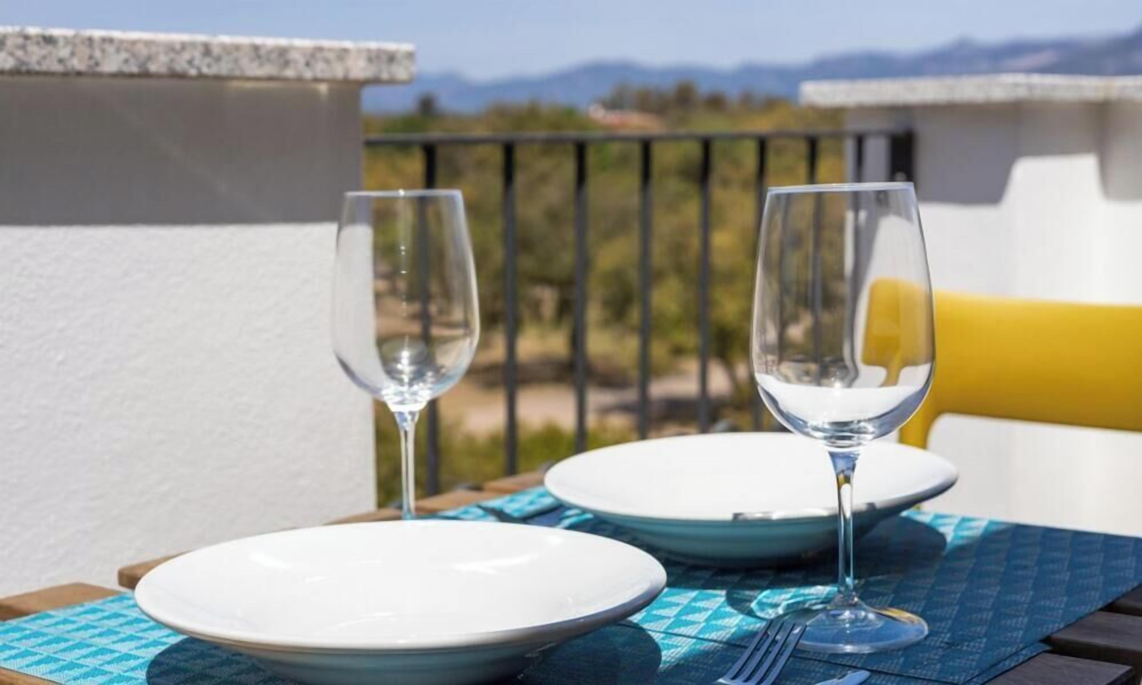 A table set with two white plates and two empty wine glasses on a blue placemat, overlooking a mountainous landscape.