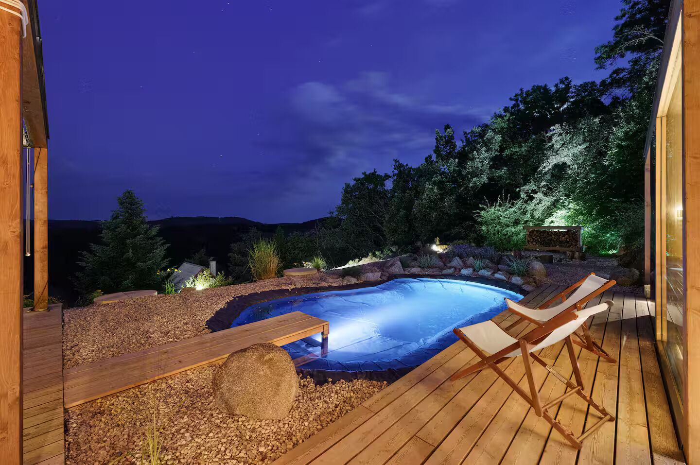 A swimming pool with wooden deck and lounge chairs, surrounded by a landscaped garden and a view of the night sky.