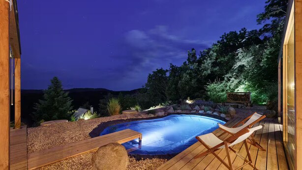 A swimming pool with wooden deck and lounge chairs, surrounded by a landscaped garden and a view of the night sky. - Sauna House Zelené Údolí (Rabyně)