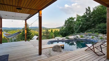 Una terraza de madera con una hamaca, un jacuzzi y sillones de descanso con vistas a un paisaje natural.