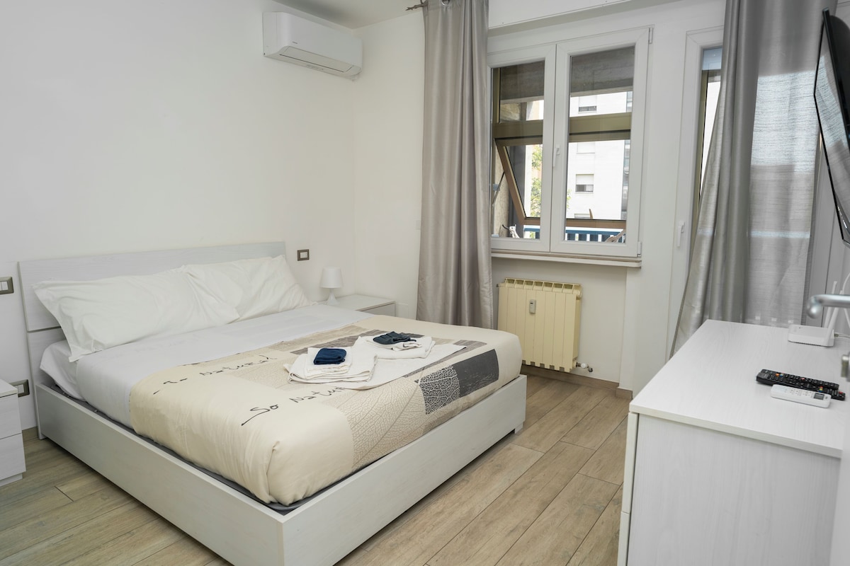 Cama bem arrumada com cabeceira branca, mesa de cabeceira, janela com cortinas e uma pequena escrivaninha com controle remoto.