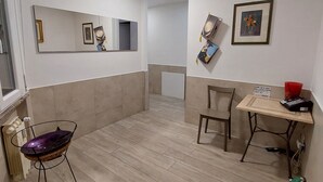 Una stanza con pavimento in legno, una sedia, un tavolino con un oggetto rosso sopra e un estintore, oltre a uno specchio appeso al muro.