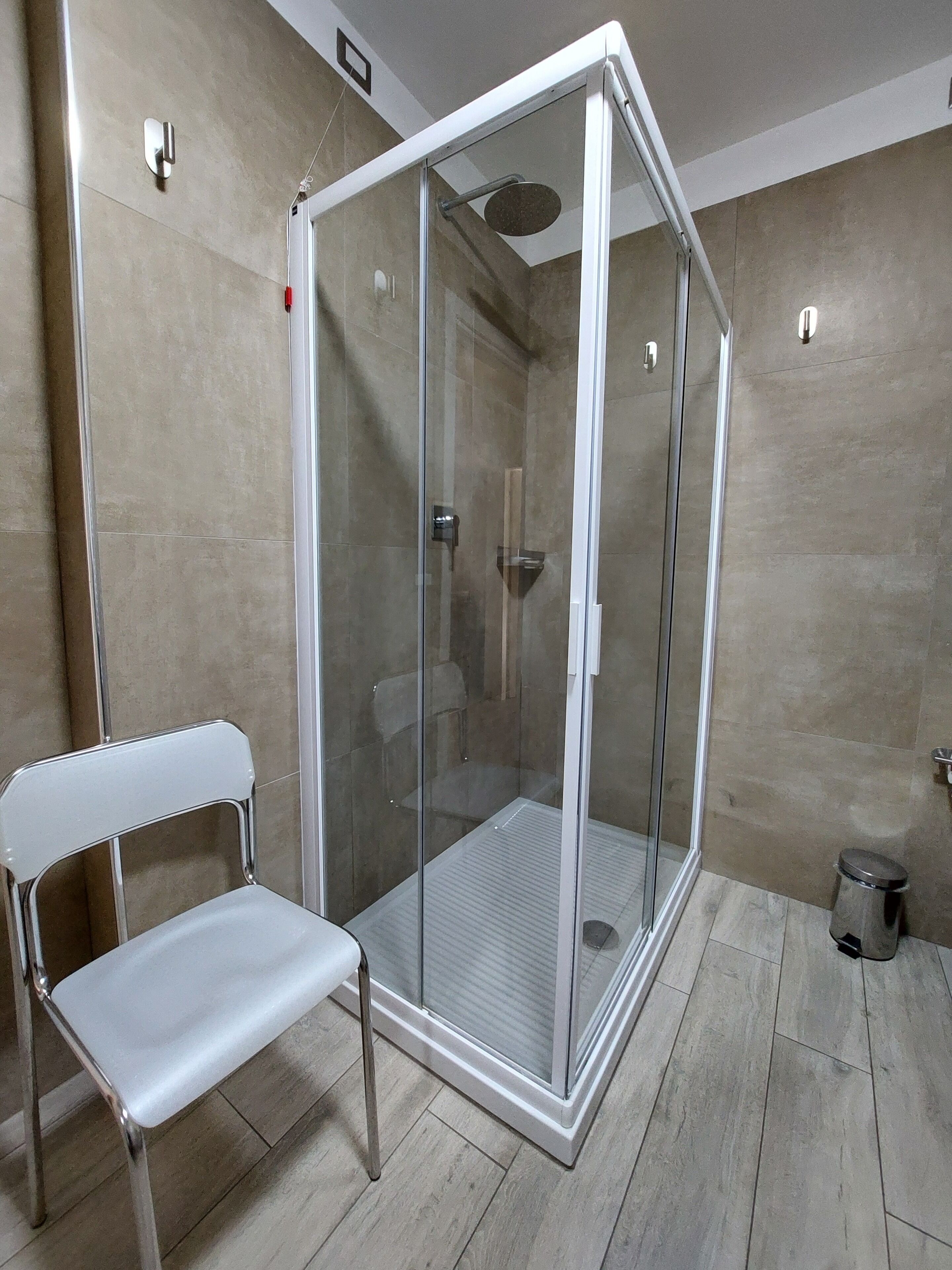 A modern shower enclosure with a glass door, a white chair, and a small trash bin.
