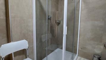 A modern shower enclosure with a glass door, a white chair, and a small trash bin.