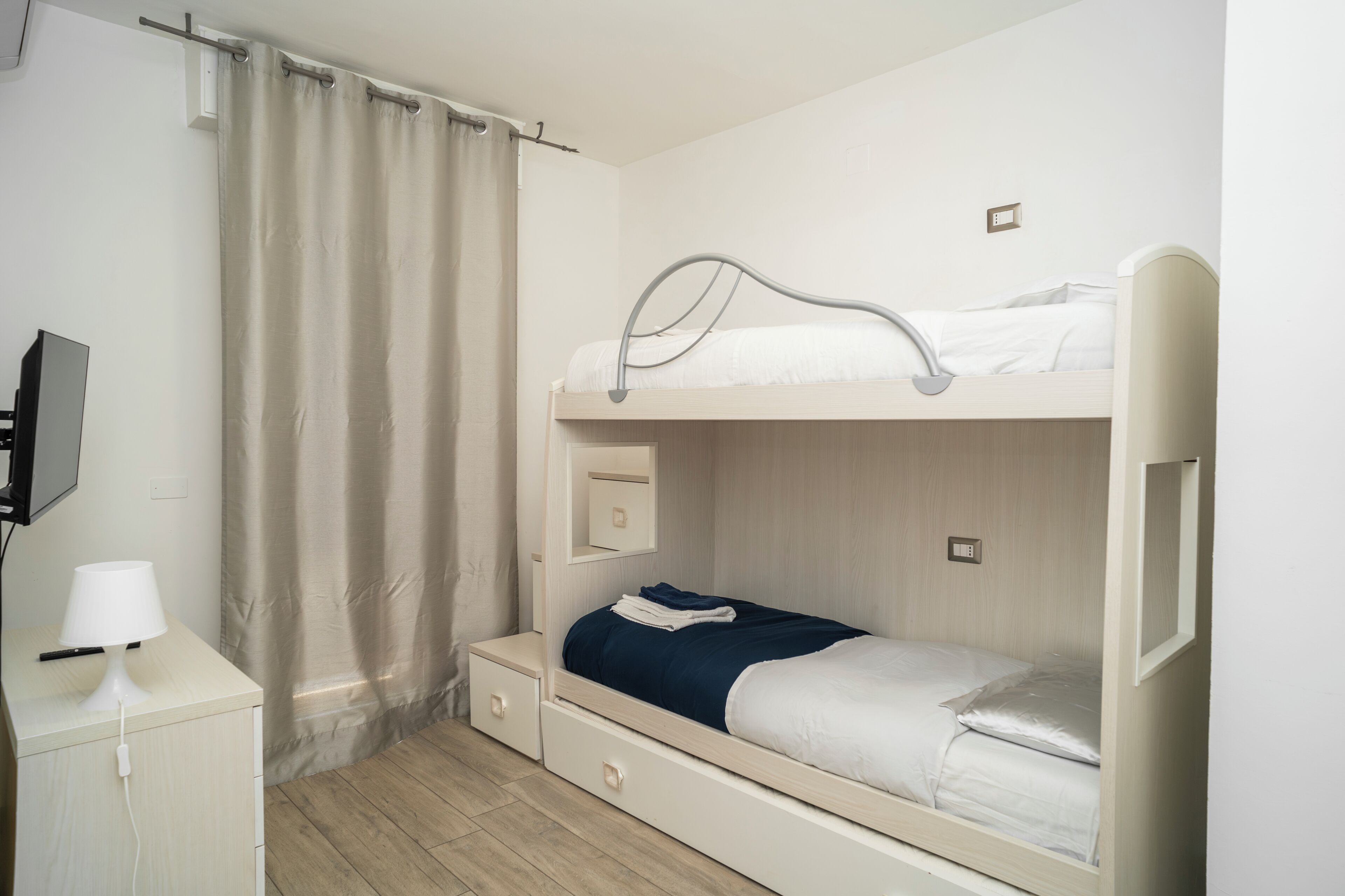 Una cama litera con escritorio, lámpara, cortina y televisor en la pared.