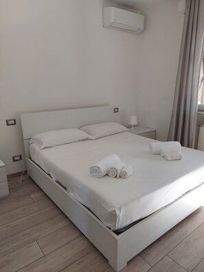 Una cama bien hecha con sábanas blancas y toallas dobladas, una mesita de noche con lámpara, y un equipo de aire acondicionado montado en la pared.