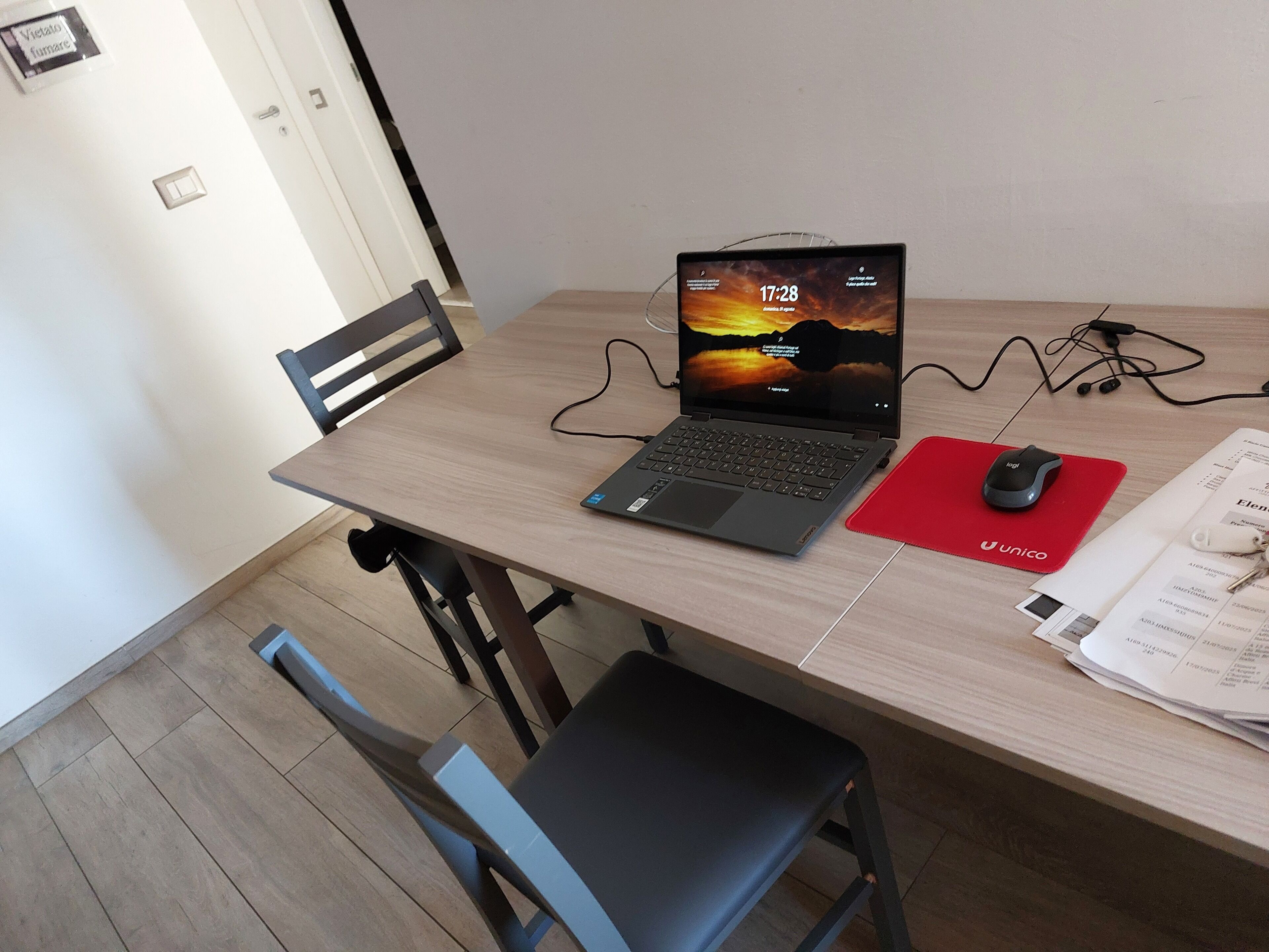 Un portatile su una scrivania di legno con un sottotavolo rosso e un mouse, una sedia e documenti sul tavolo.