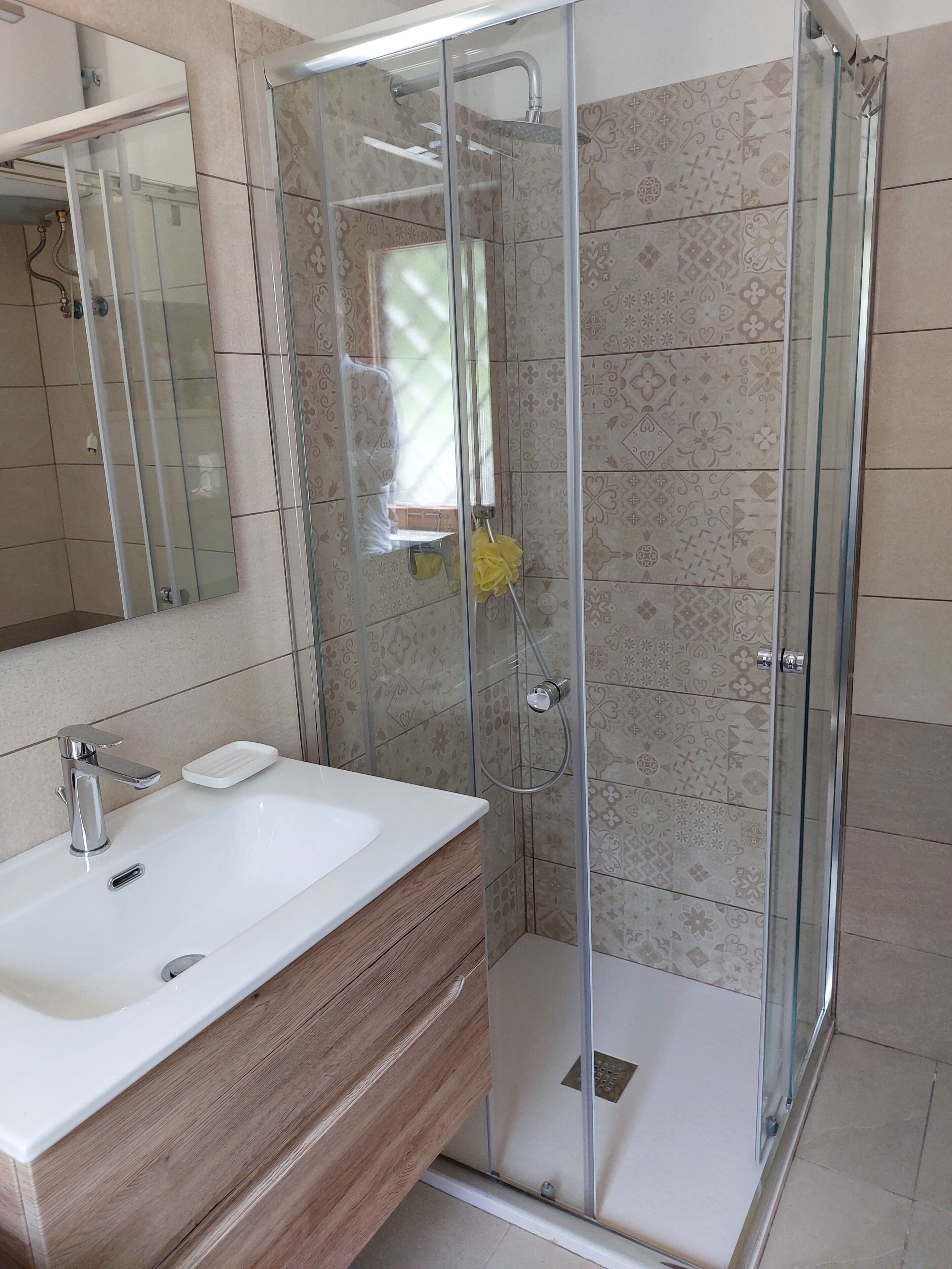 Une salle de bain dotée d’une douche à parois de verre, d’un lavabo blanc avec un meuble-vasque en bois et de carreaux décoratifs sur les murs.