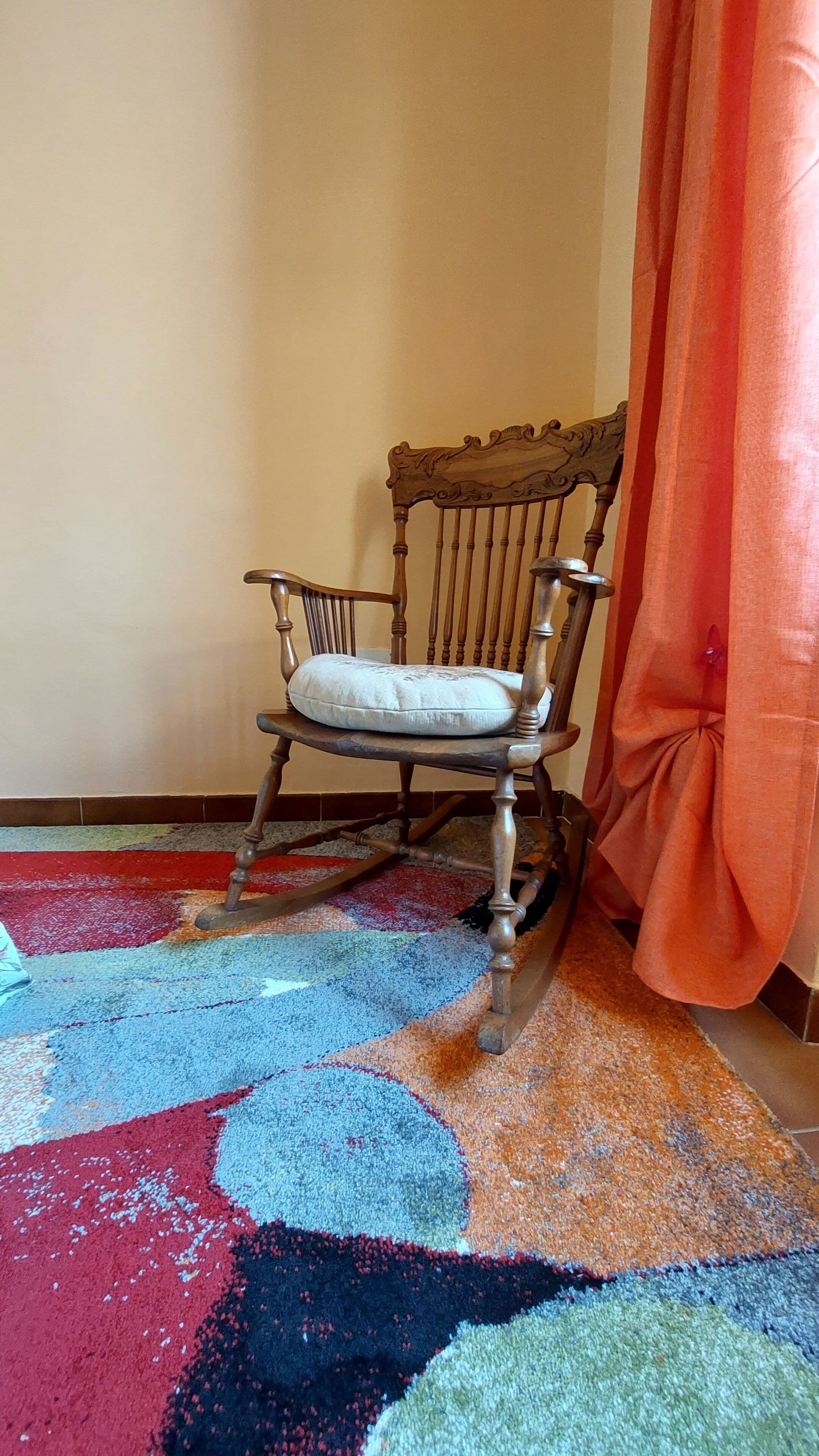 A rocking chair with a cushion, a colorful rug, and a red curtain.