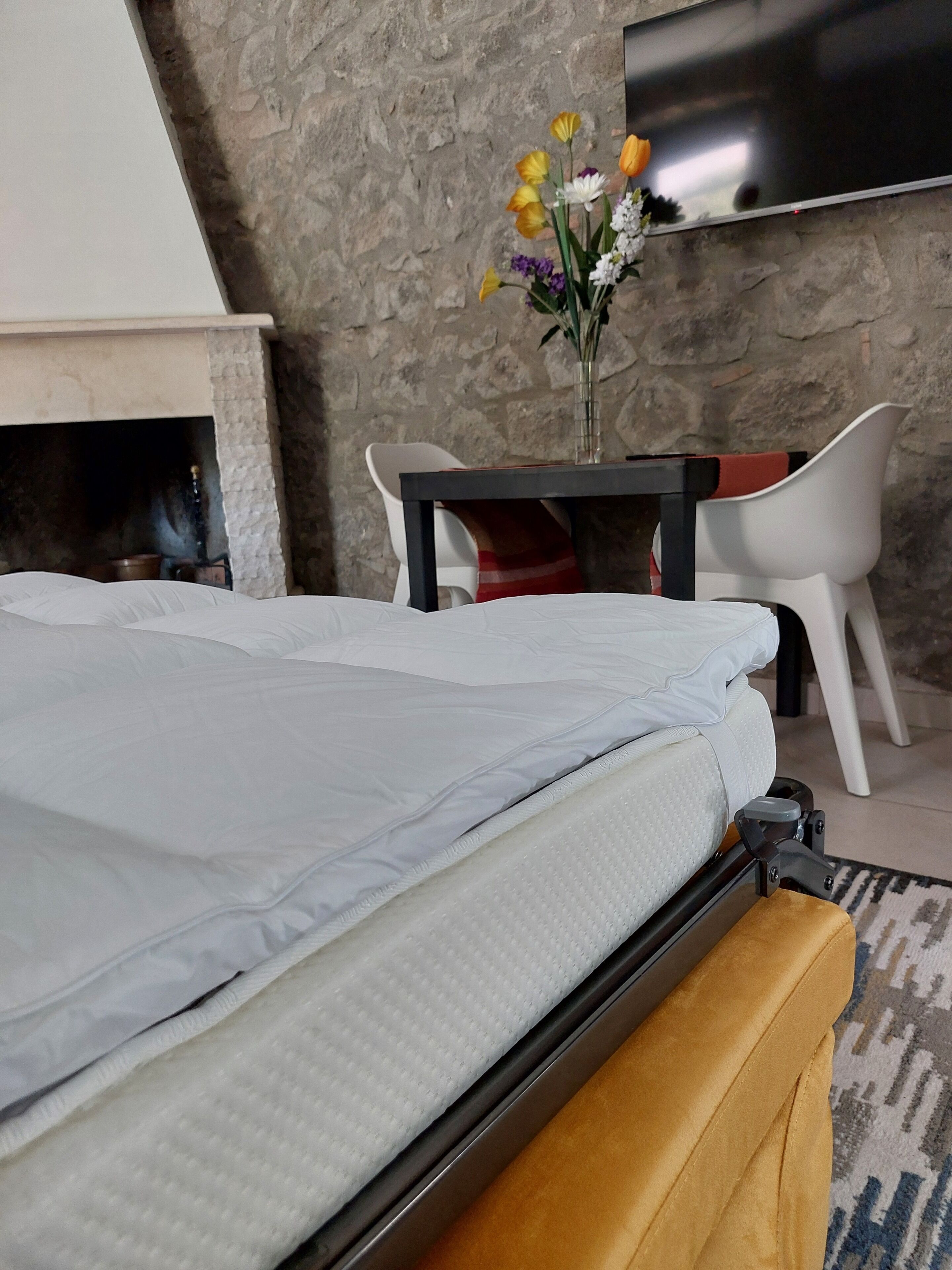 Une chambre à coucher avec un lit, un téléviseur fixé au mur, un mur en pierre, une cheminée et une table avec des chaises.