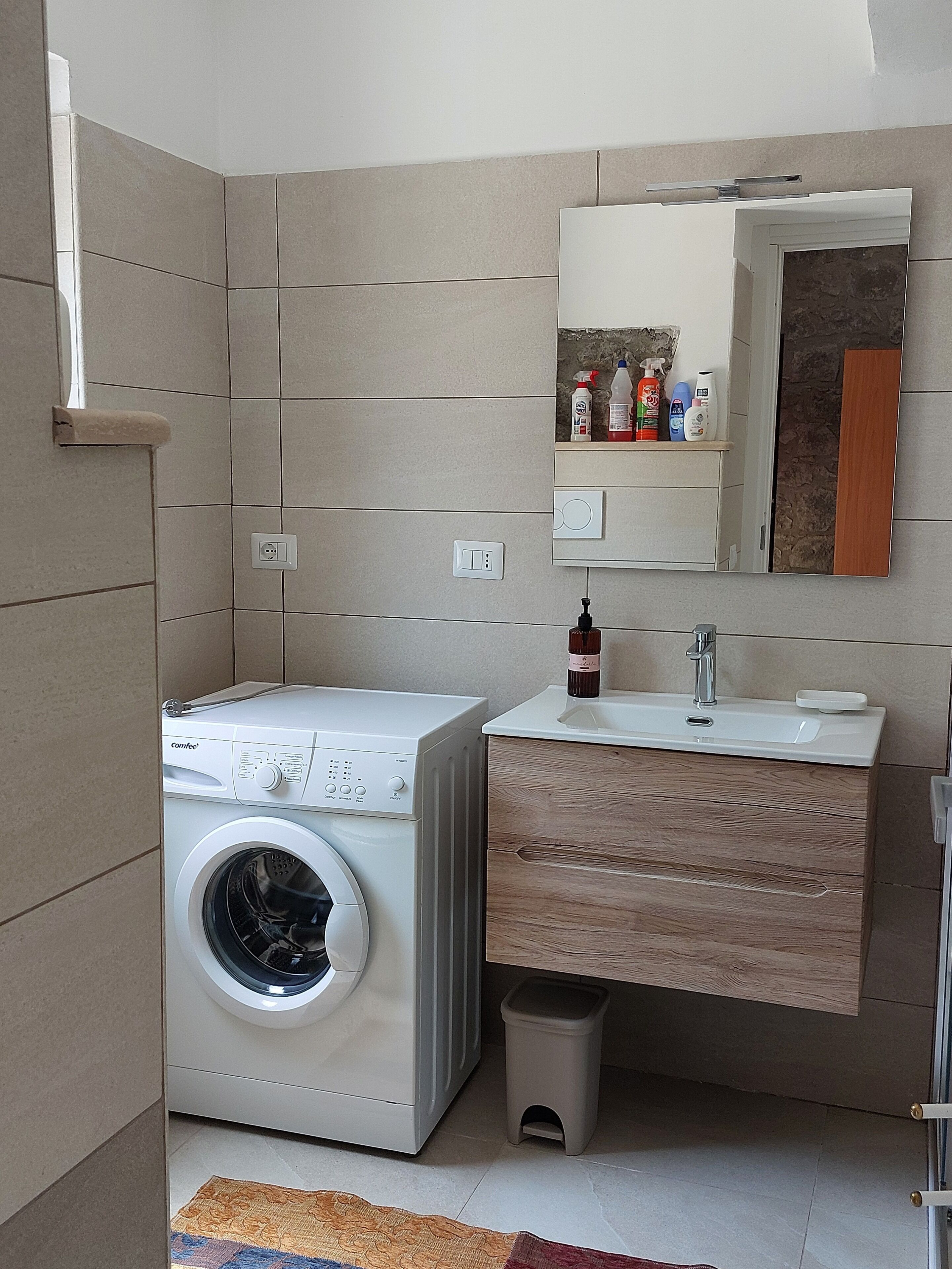 A compact laundry area with a front-loading washing machine, a sink with a mirror above, and a small trash bin.