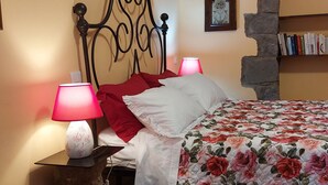 Un dormitorio con cama, dos lámparas de noche, almohada roja, colcha con estampado floral, pared de piedra, estantería y alfombra estampada.