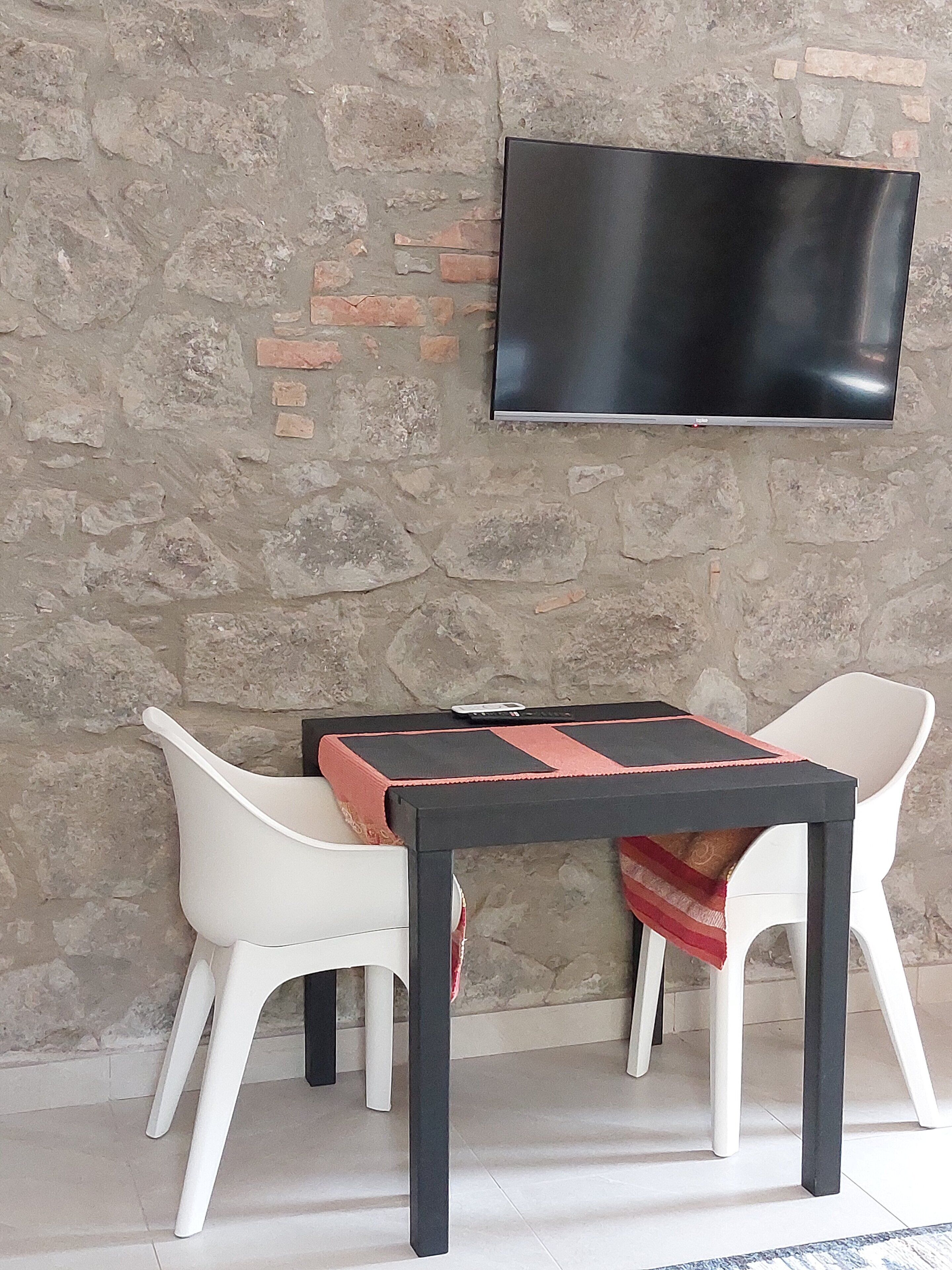 Une pièce avec un mur en pierre, un téléviseur à écran plat fixé au-dessus d’une table, et deux chaises blanches.