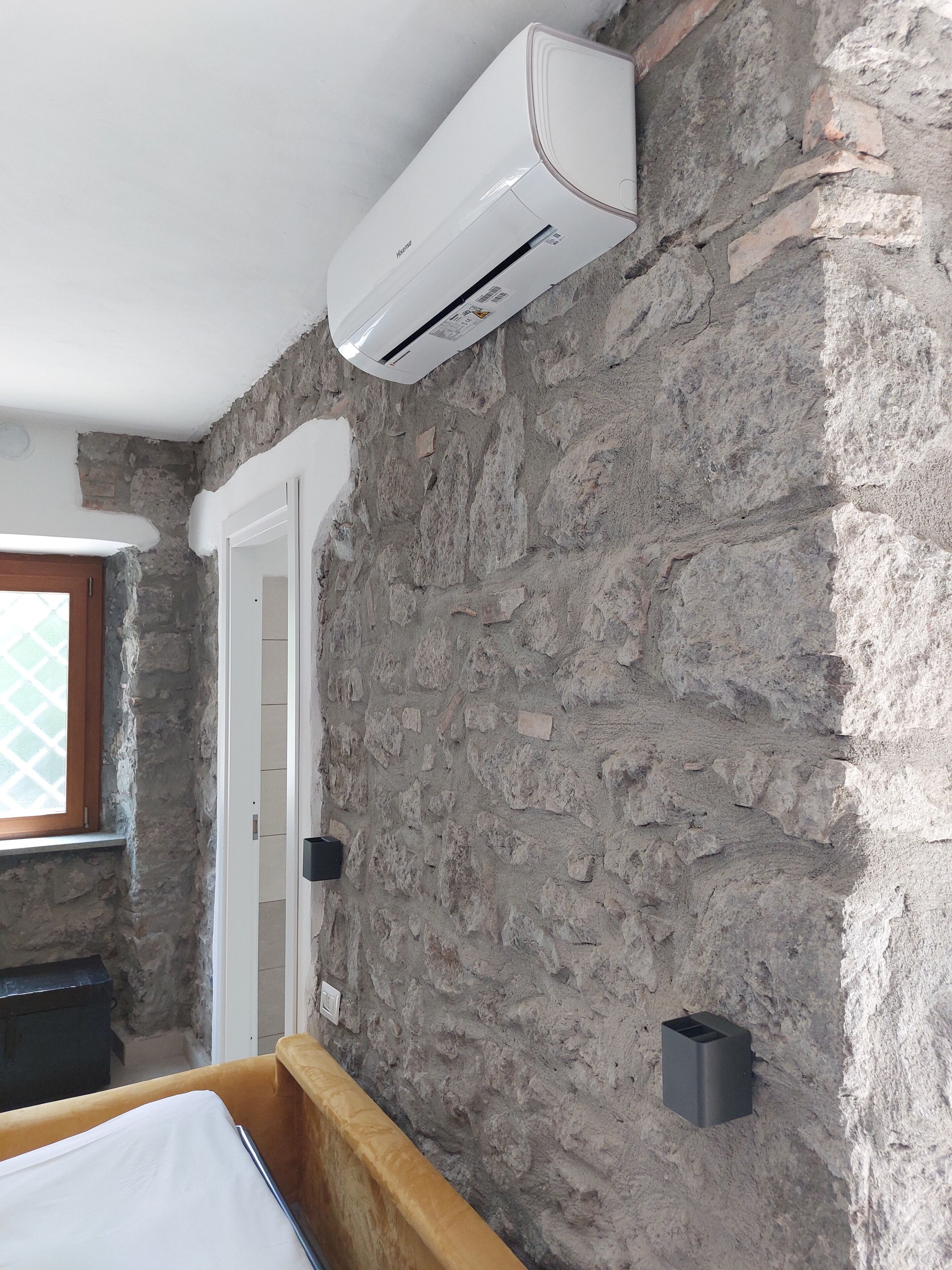 Une pièce avec un mur en pierre, un lit, un climatiseur blanc et un petit objet noir accroché au mur.