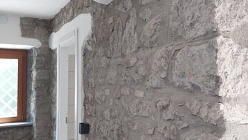 Une pièce avec un mur en pierre, un lit, un climatiseur blanc et un petit objet noir accroché au mur.
