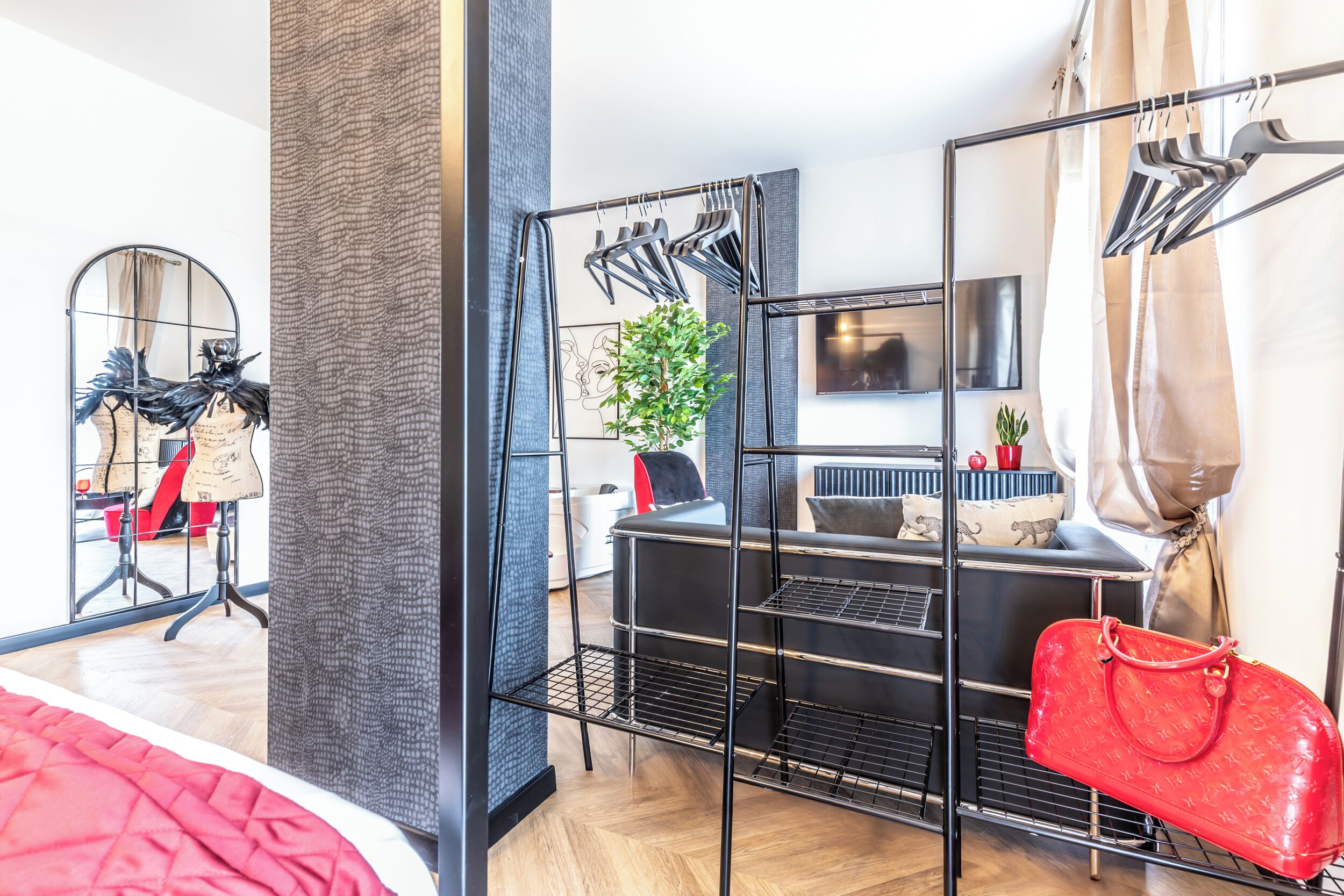 A modern hotel room with a black metal rack, a red handbag, a plant, and a TV mounted on the wall.