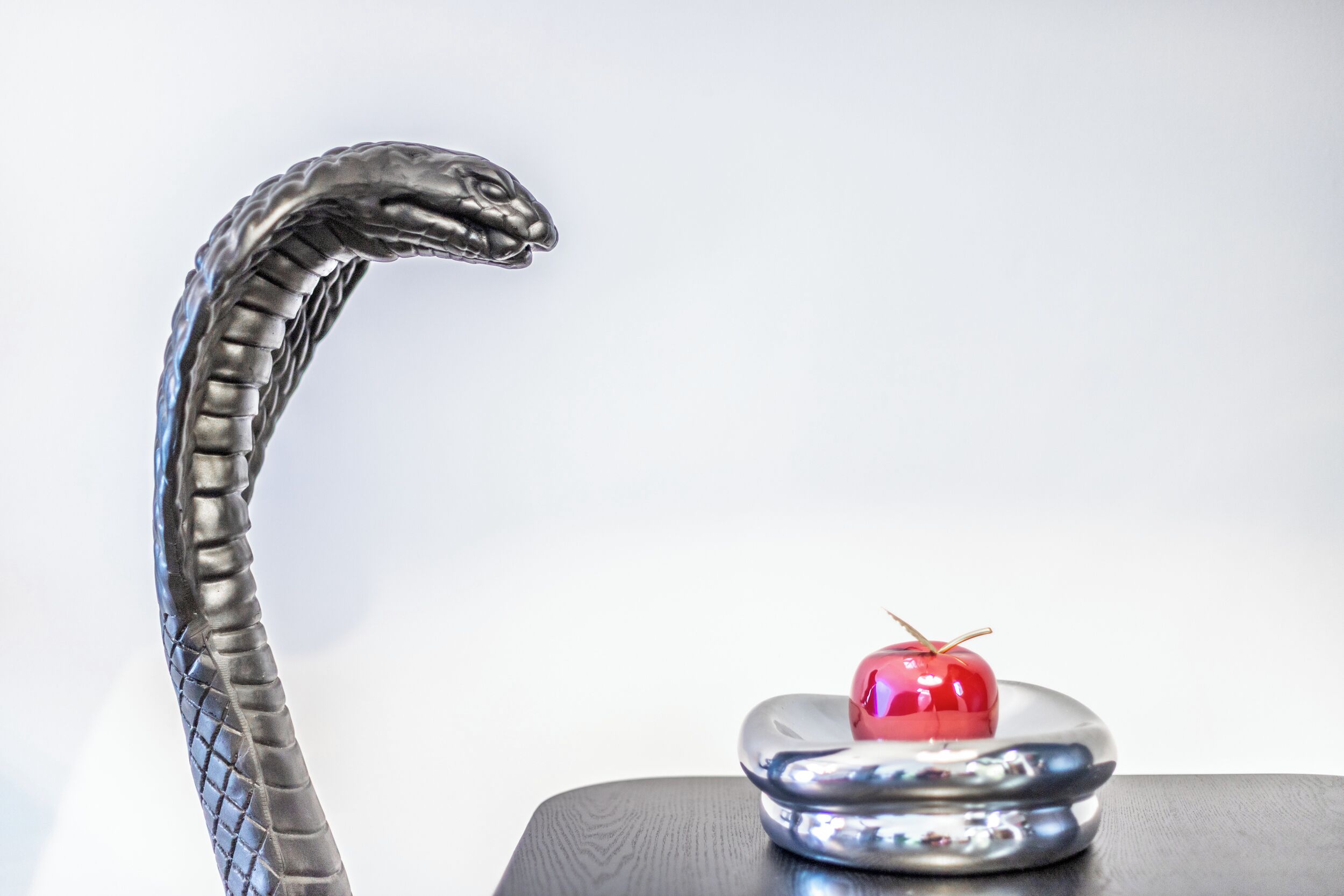 Una escultura metálica de serpiente con una manzana roja sobre una superficie reflectante.