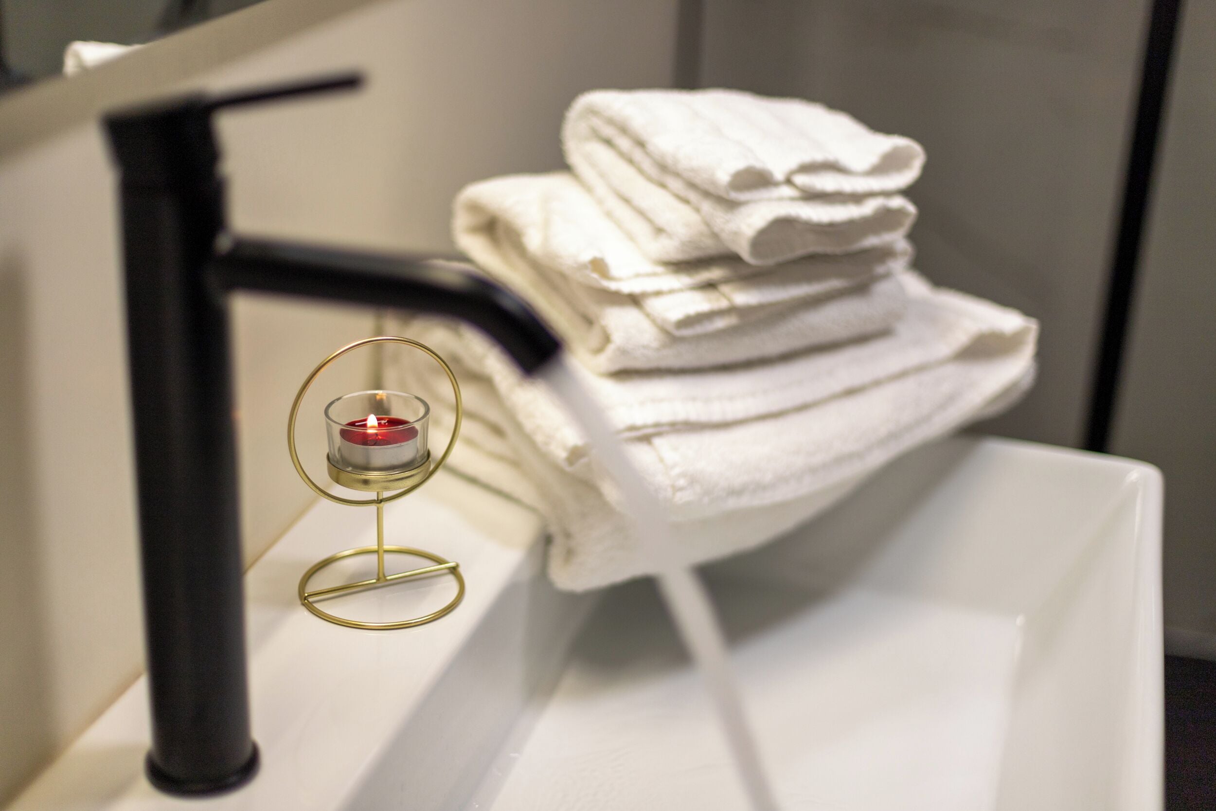 Une salle de bain avec une pile de serviettes blanches sur une étagère blanche, un robinet noir et un porte-bougie avec une bougie allumée.