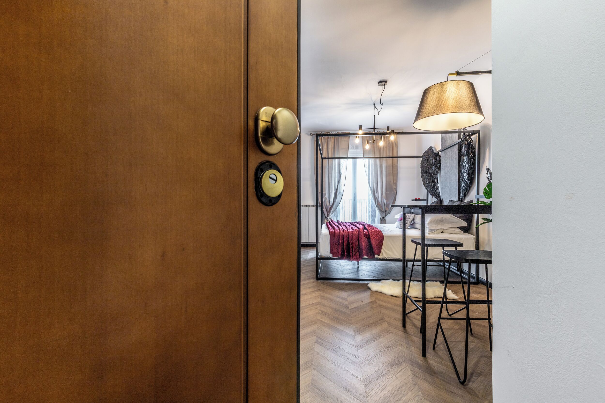 Moderni hotellihuone, jossa on puinen ovi, punaisella peitolla varustettu sänky, musta metallinen työpöytä ja suuri lamppu.