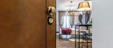 A modern hotel room with a wooden door, a bed with a red blanket, a black metal desk, and a large lamp.