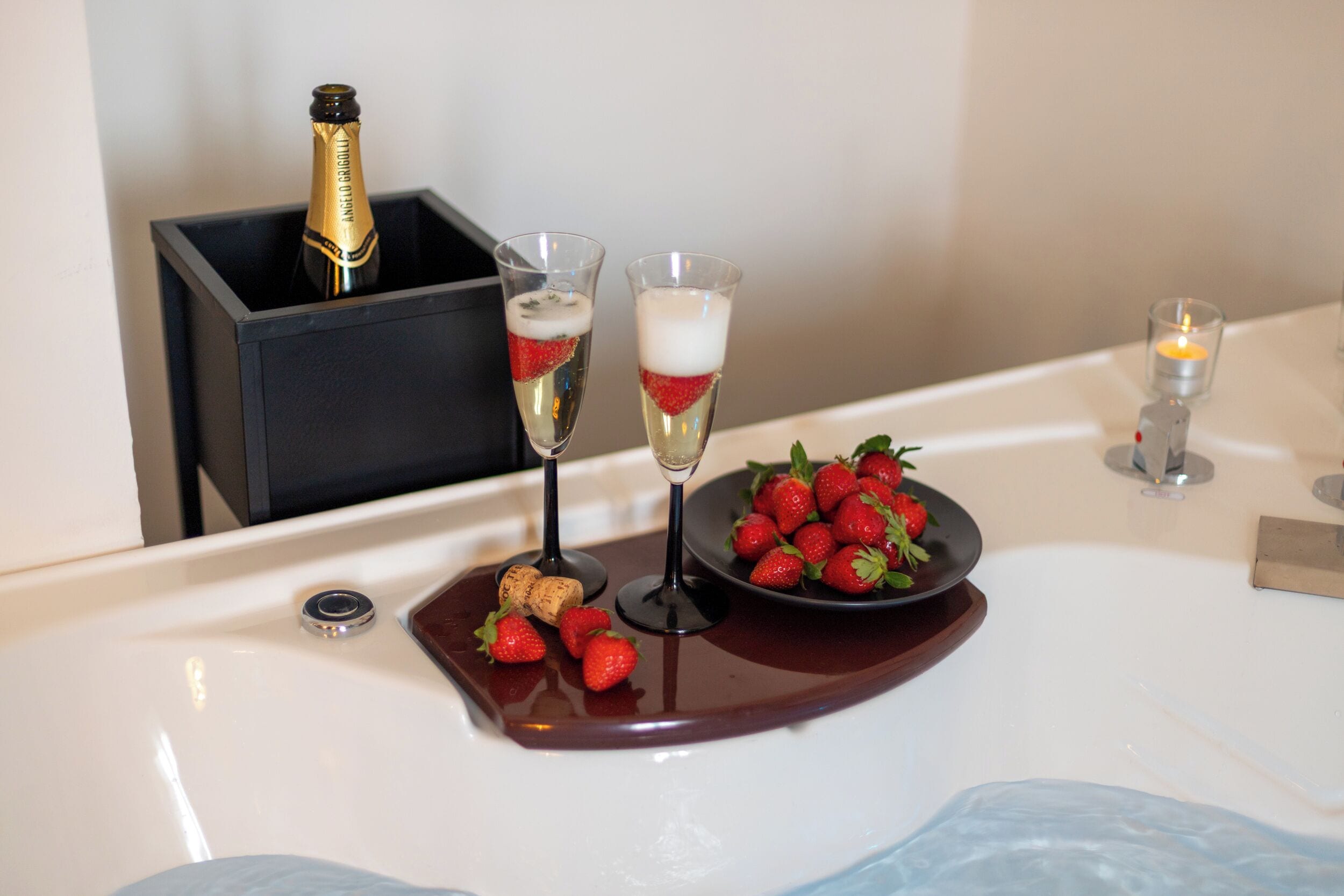 Una botella de champán en una caja negra, dos copas de champán con fresas, un cuenco de fresas y una vela sobre una mesa blanca.