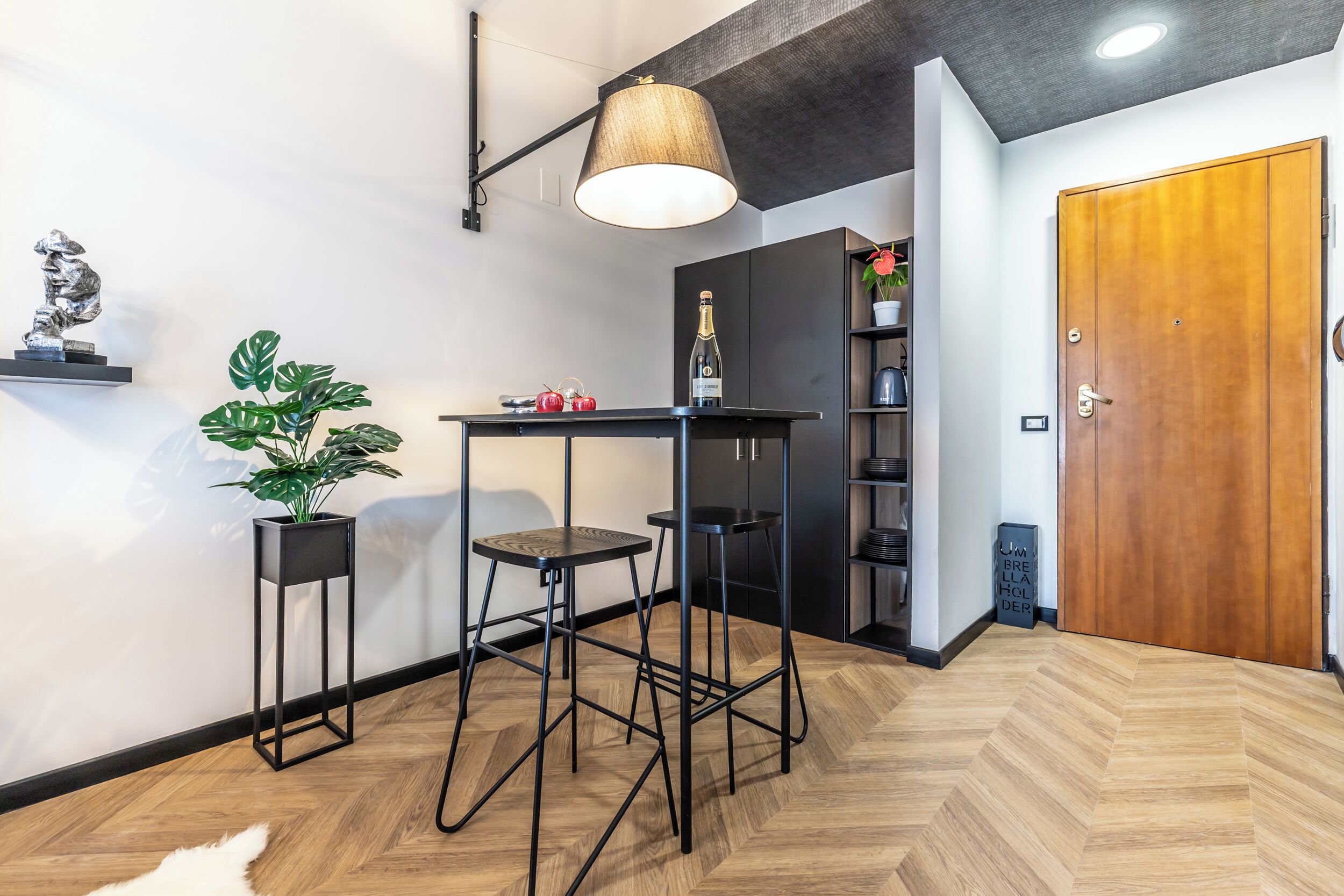 Un interno moderno con pavimento in parquet a spina di pesce, un bancone in metallo nero con sgabelli, una lampada a sospensione e una porta in legno.