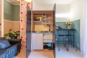 Uma cozinha compacta equipada com micro-ondas, prateleiras e um pequeno frigorífico. Há um banco de bar e um vaso com planta no quarto.