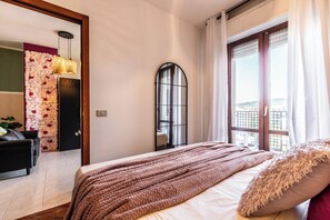 A bedroom with a bed, a window with curtains, a mirror, and a wall with a floral arrangement. - Luxury Suite - Tentazioni Romantiche (Perugia)