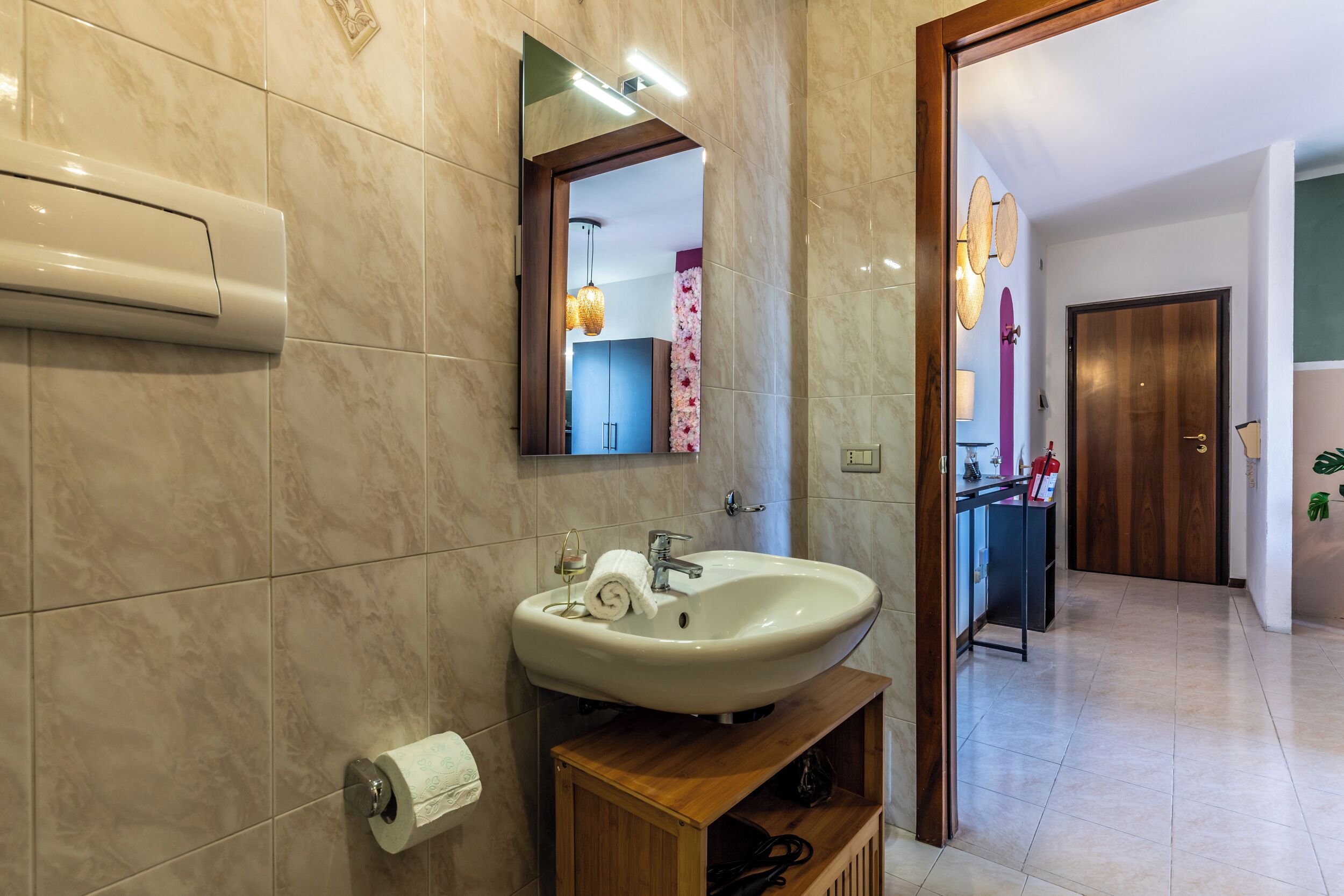 A modern bathroom with a marble-tiled wall, a white sink, a mirror, and a wooden shelf.