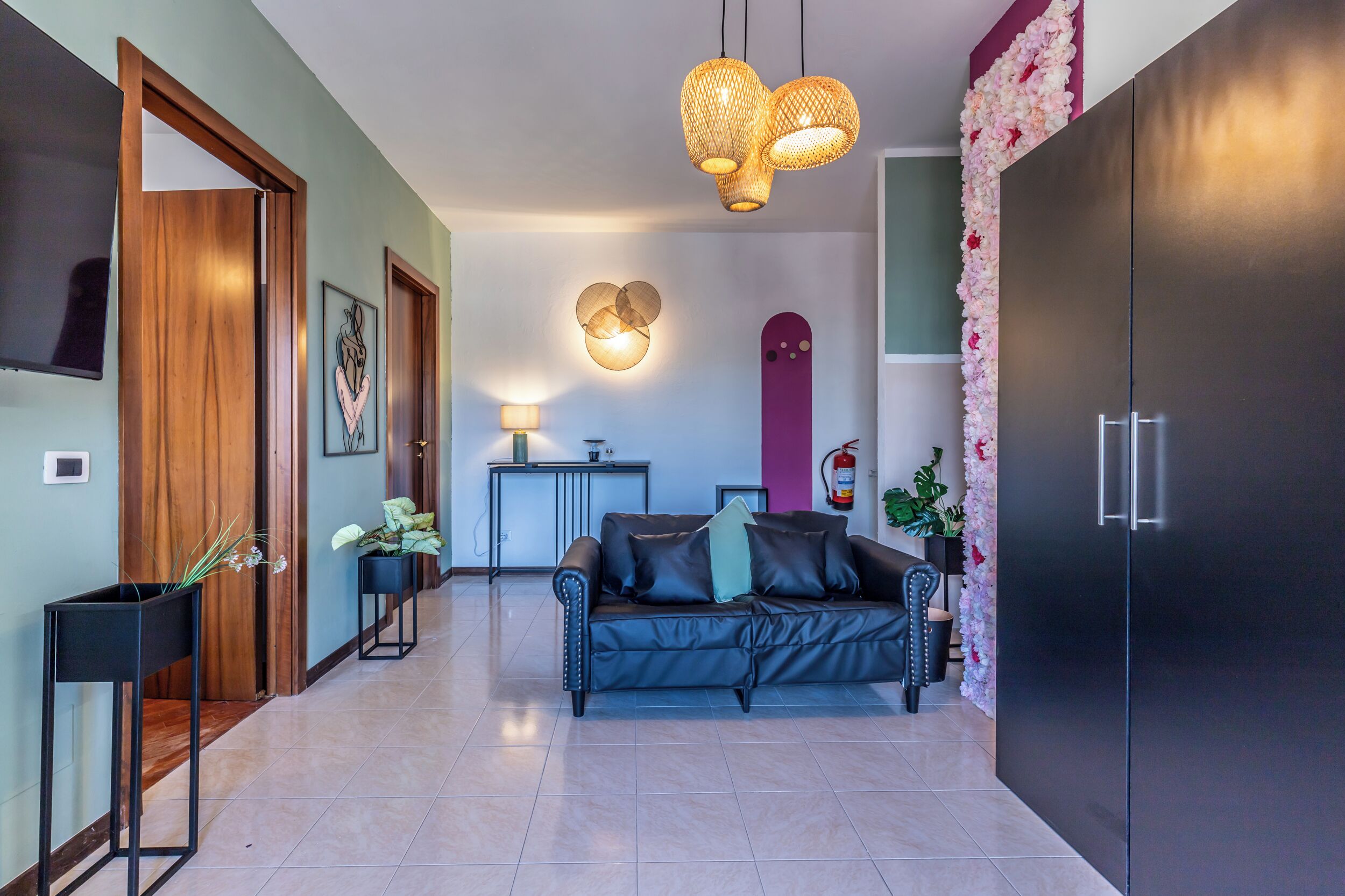 Ein modernes Wohnzimmer mit einem schwarzen Ledersofa, einer Holztür, einem schwarzen Schrank, einem an der Wand montierten Fernseher und einer Wand mit Blumen als Dekoration.