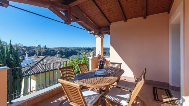 A wooden balcony with a table and chairs, overlooking a scenic view of houses and trees. - A 15 Miglia da Roma (Riano)