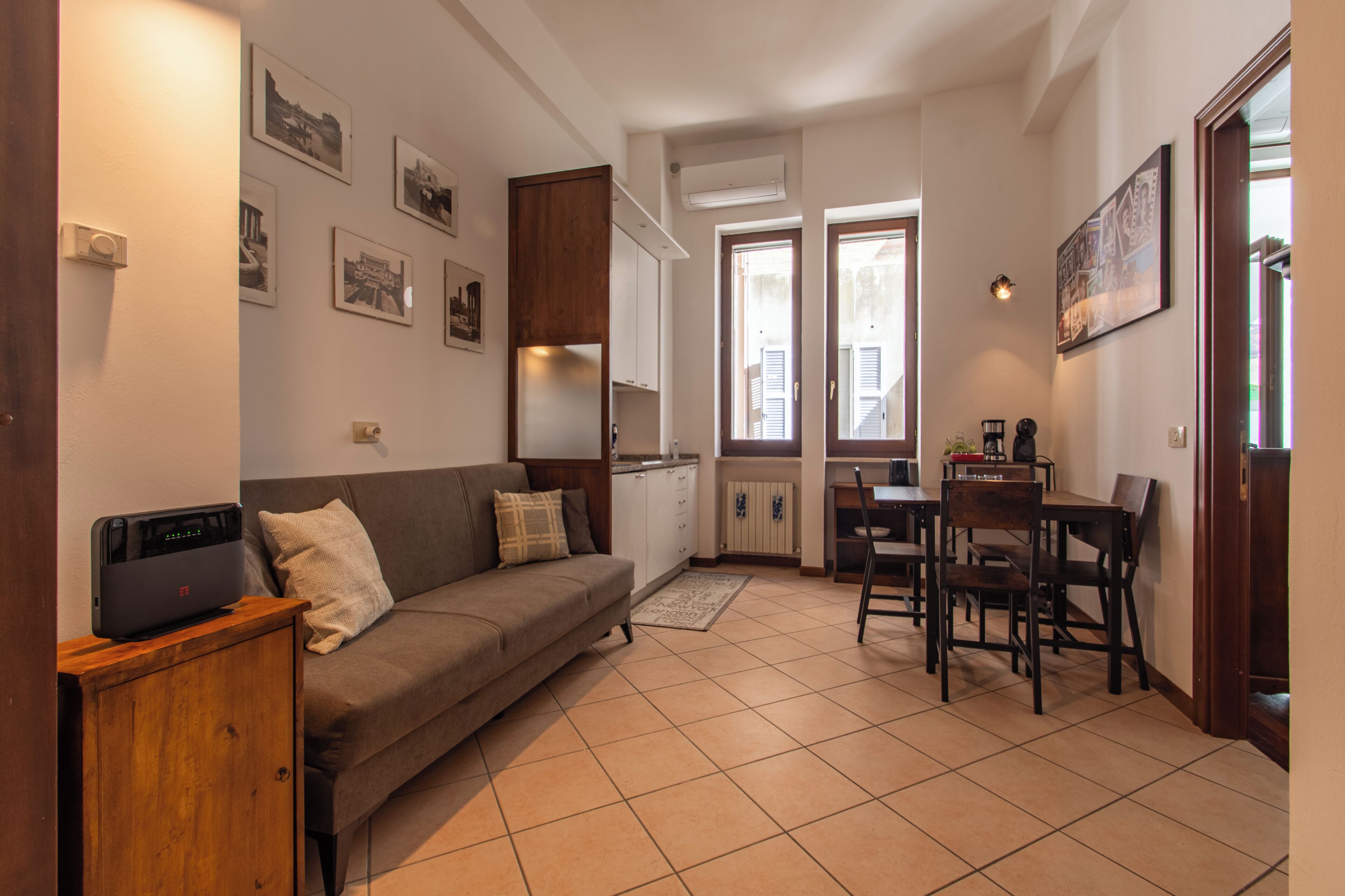A compact living space with a sofa, dining table, and chairs. There are framed pictures on the walls, a cabinet, and a radiator.