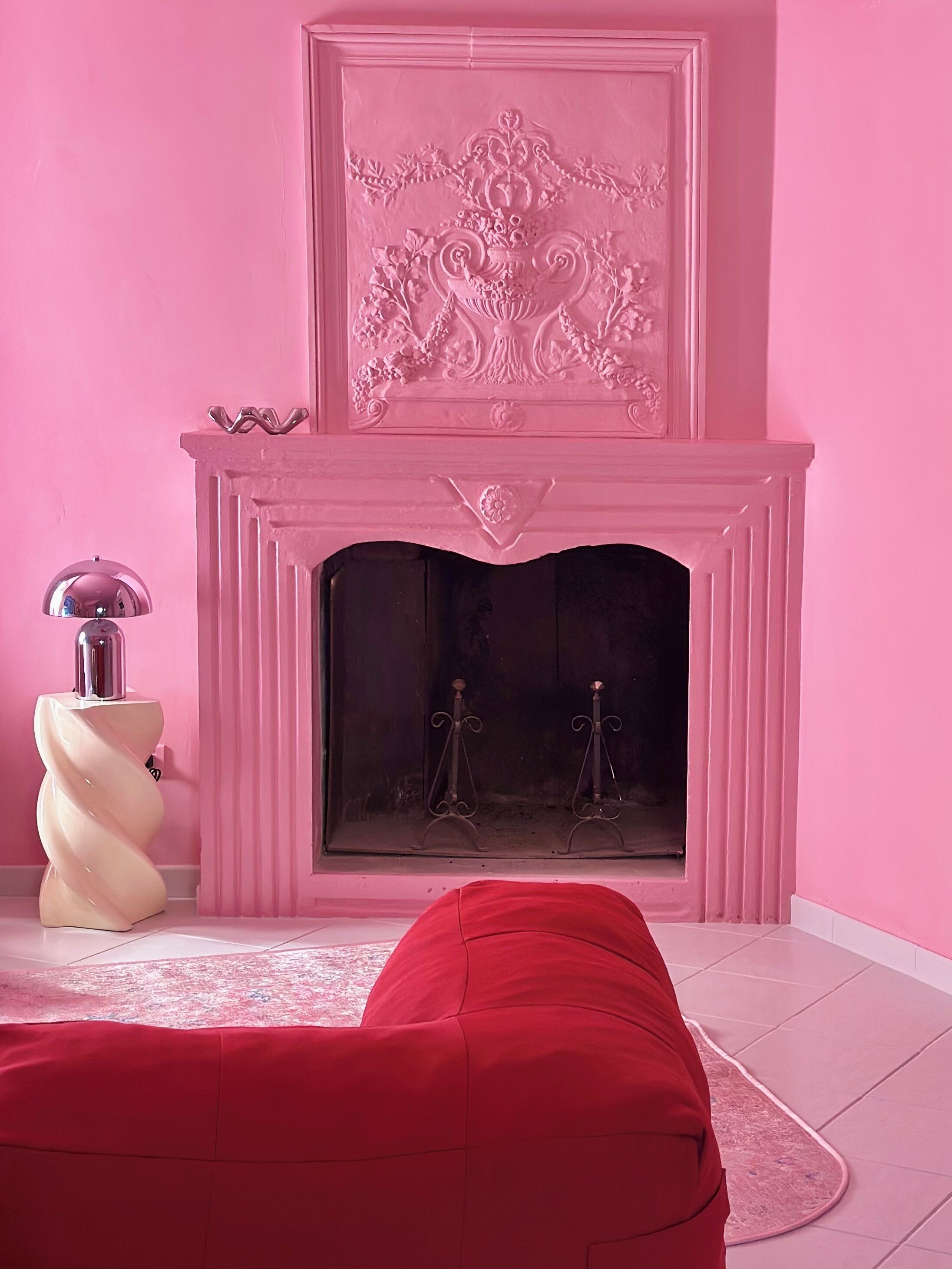 A room with a pink wall, a fireplace with a decorative panel above it, a red sofa, and a white side table with a lamp.