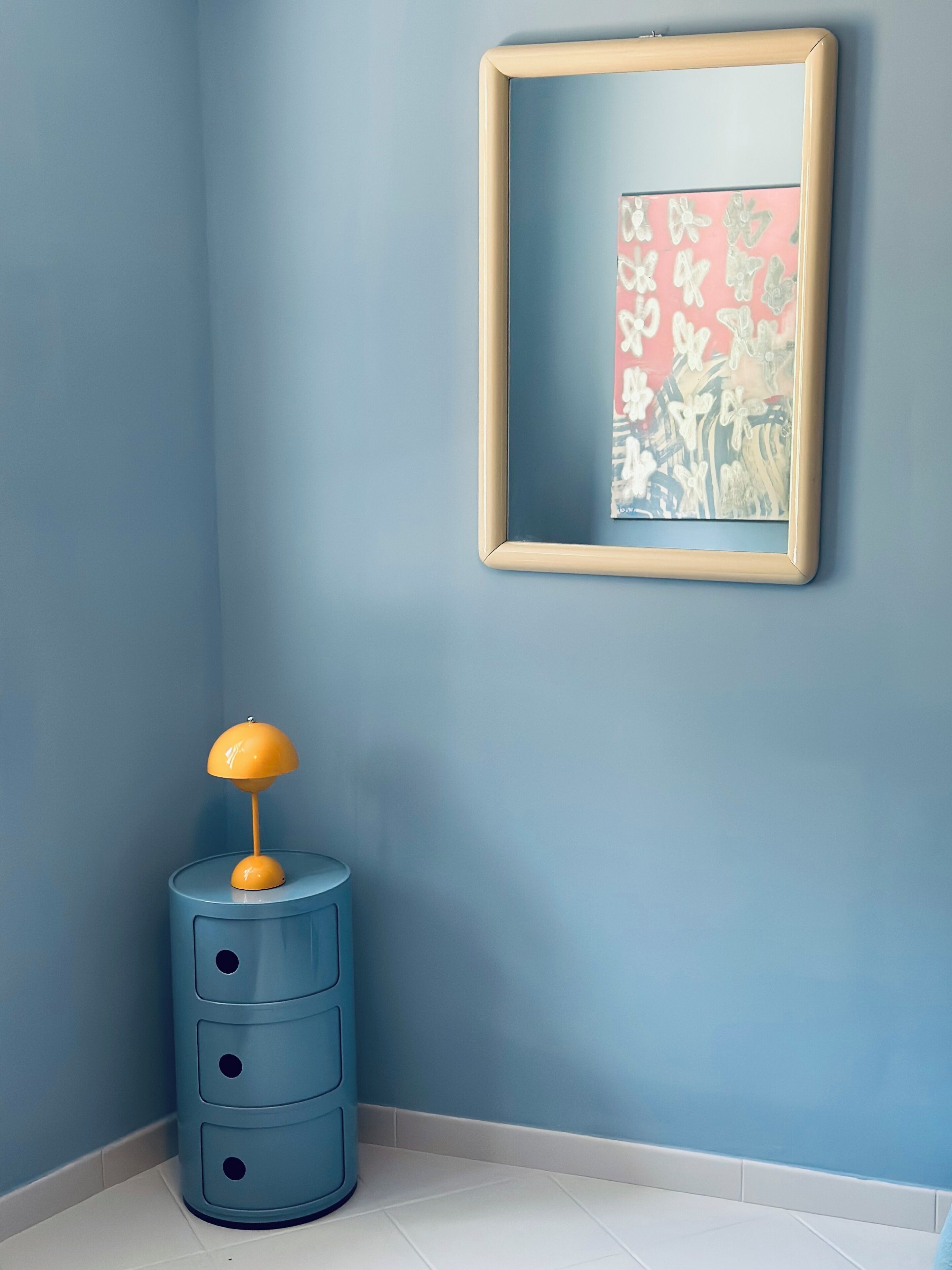 A corner room with a blue wall, a framed artwork, a blue side table with a yellow lamp, and a white floor.