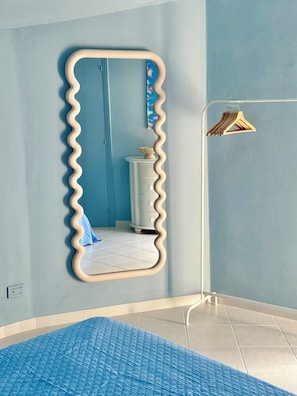 Una stanza con uno specchio ondulato, una cassettiera bianca, un tappeto blu e un appendiabiti con grucce.