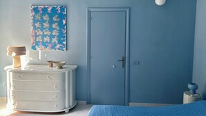 A bedroom with a blue door, a white dresser, a blue bedspread, and a wall-mounted lamp.