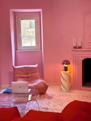 A pink-themed room with a bed, a laptop, a lamp, and a fireplace.