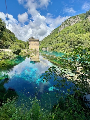 Una veduta chiara di un fiume con una struttura in pietra al centro, circondata da vegetazione rigogliosa e un panorama montuoso.