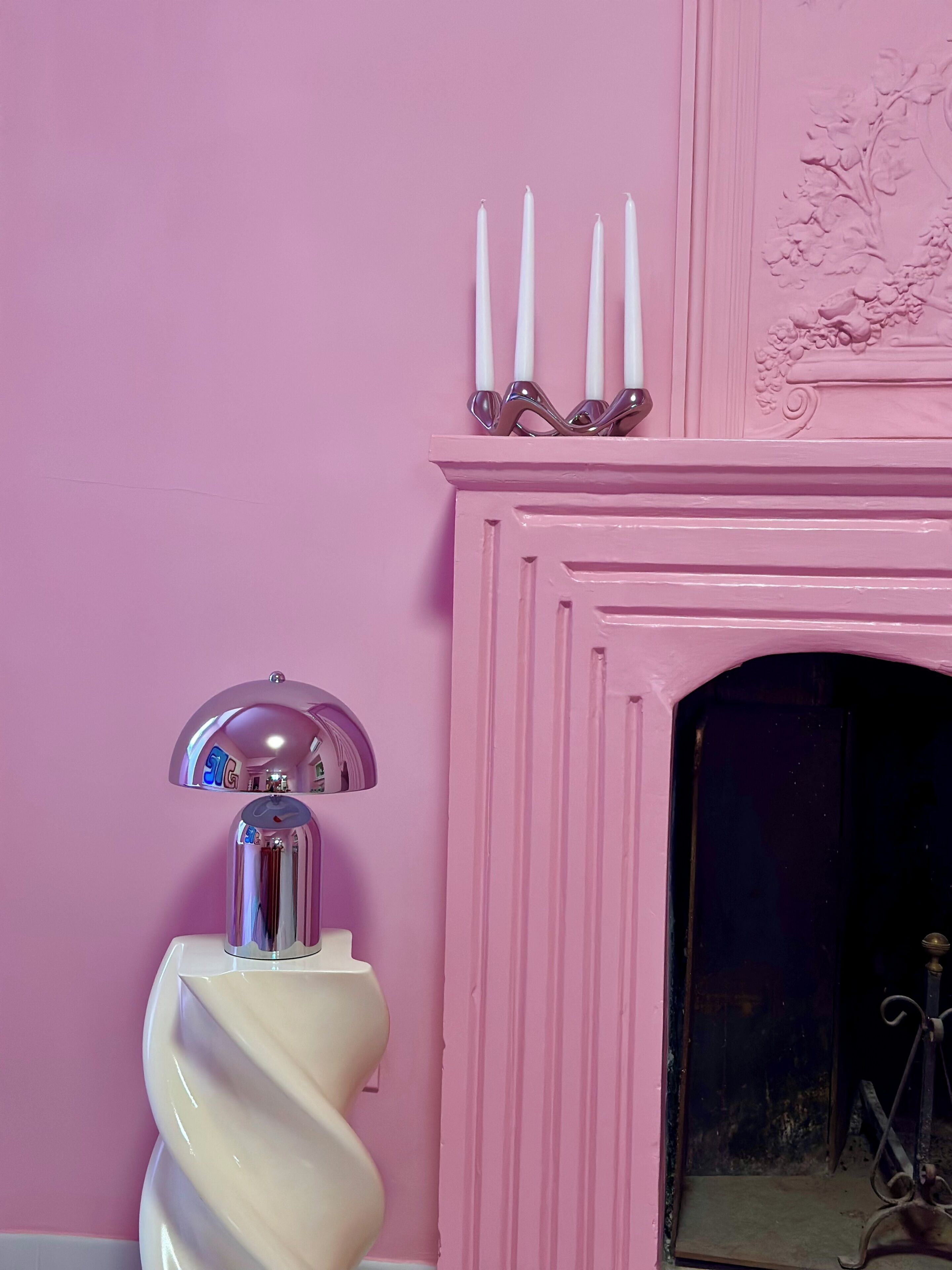 A pink wall with a white fireplace, three white candles, and a modern lamp.