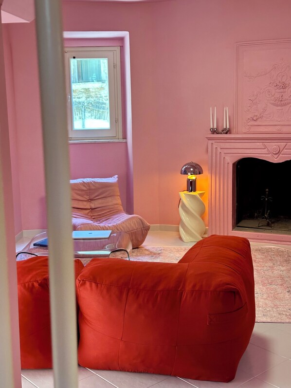 A pink-themed room with a bed, a red bean bag, a lamp, and a fireplace.