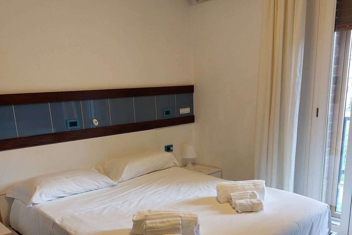 Habitación de hotel con cama, mesitas de noche, aire acondicionado y una ventana con cortinas.