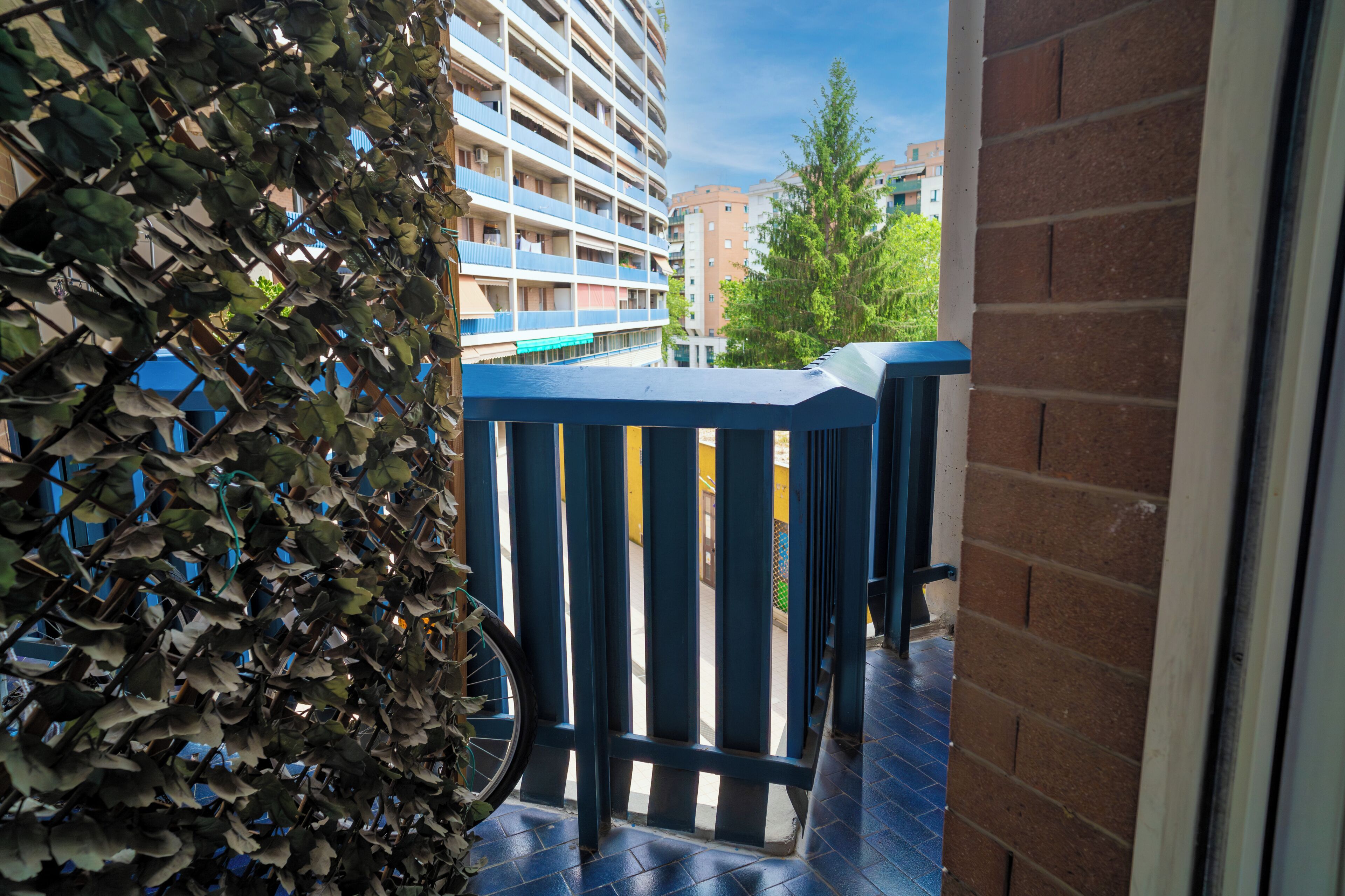 A balcony with a blue railing, a bicycle, and a view of a multi-story building in the background.