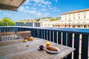 Una mesa de madera con artículos de desayuno, como un croissant, una taza de café y un vaso de zumo de naranja, ubicada en un balcón con vistas a edificios y árboles.