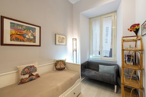 A cozy room with a bed, a grey sofa, a window with curtains, a bookshelf, and a framed painting on the wall. - A 20 Gradini dai Musei Vaticani (Rome)