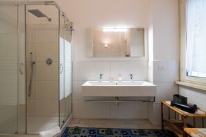 Ein modernes Badezimmer mit einer ein Glasabdeckung umfassenden Dusche, einem weißen Waschbecken mit Spiegel darüber und einem kleinen Holztisch mit einem Korb.