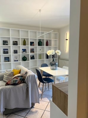 A modern living room with a white sofa, a dining table, and a bookshelf filled with various items.
