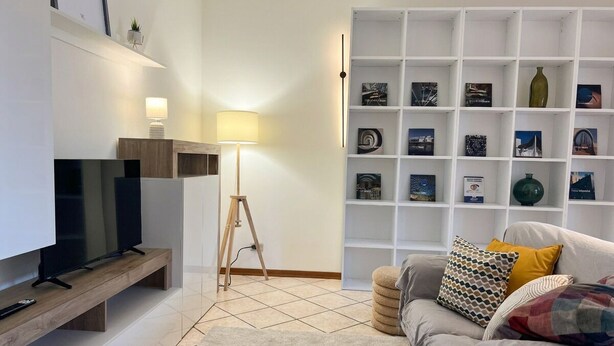 A living room with a TV on a wooden stand, a lamp, a bookshelf, and a sofa with cushions. - Tentazioni Raffinate - Luxury Suite (Perugia)